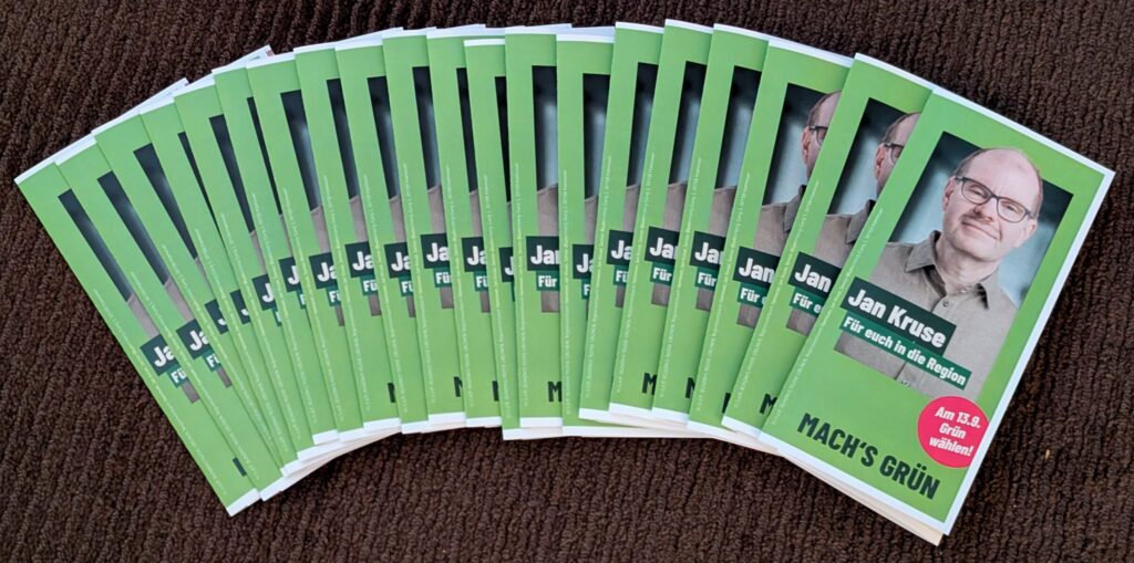 Auf einem dunkelbraunen Teppich liegen 20 Wahlkampfflyer aufgefächert. Es ist dir grüne Vorderseite zu sehen mit einen Bild einer männlichen Person. Text über dem Bild: "Jan Kruse" "Für euch in die Region" Unten der Claim "MACH'S GRÜN" Außerdem ein pinkfarbener Kreis, in dem "Am 13.9.Grünm wählen!"