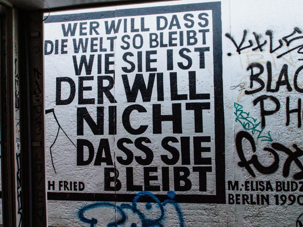 Schwarzer Schriftzug auf weißer Wand in Form eines mit Schablone aufgetragenen Graffito: "WER WILL DASS DIE WELT SO BLEIBT WIE SIE IST DER WILL NICHT DASS SIE BLEIBT"!