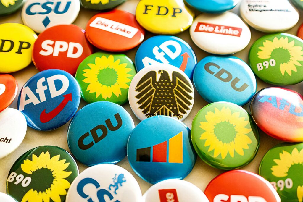 Eine Ansammlung von Button-Pins mit den Logos verschiedener Parteien. Ein Button-Pin trägt den Bundesadler.